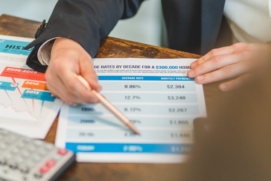 A person in formal attire is seated at a wooden table, pointing to a printed chart displaying home mortgage interest rates by decade, with figures such as average rates and monthly payments. Several o