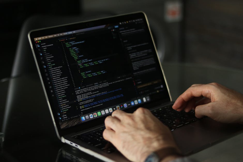 A person working on a laptop computer with a dark-themed code editor visible on the screen, situated on a black table. The individual's hands are positioned over the keyboard, suggesting active typing
